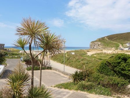 A coastal view with palm trees a parking area a road sandy beach grassy hills and a cliff at Kingsholm in Porthtowan