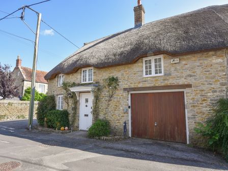 A stone cottage with a thatched roof a white door and wooden garage door on a street at Stable Cottage in Burton Bradstock