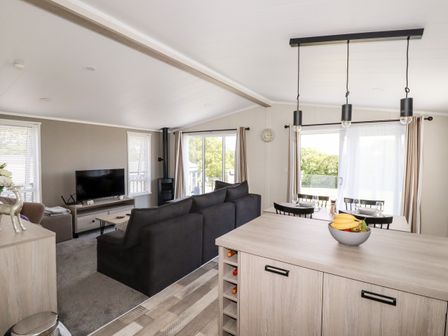 An open plan living and dining area with a black sofa a television a dining table set for four and a kitchen island with fruit bowl at 63 The Maples in Hewas Water near Sticker