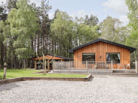 A wooden cabin with a deck and metal chairs surrounded by trees and a gravel area at Lovat Highland Bothy in Beauly