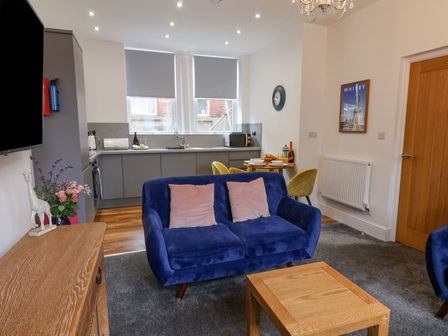 A living room with a blue sofa and pink cushions a wooden coffee table wooden cabinet and an open kitchen with grey cabinets and dining table at Routh Retreat Whitby
