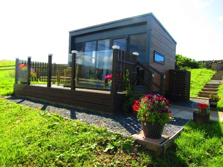 A small modern cabin with glass railing and flower pots in a grassy area at Balvicar Beag in Balvicar Isle Of Seil