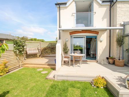 A garden with grass a wooden deck with a swing and a patio with chairs and a table at Blue Waters in Weymouth