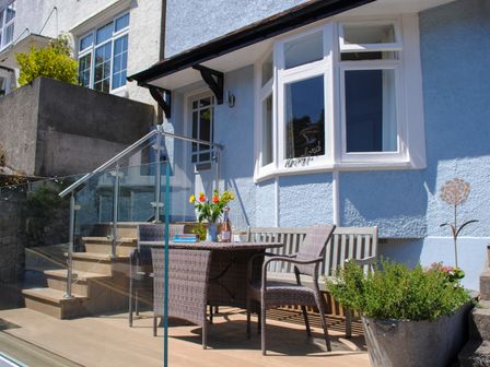 An outdoor patio area with a wicker table and chairs flowers on the table glass railing and blue house exterior at Cranford in Salcombe