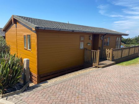 A wooden cabin with a small porch and railing next to a paved pathway at Mojo Mullacott near Ilfracombe