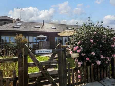 A wooden gate with a sign reading Fern Cottage a bush with pink flowers and a house with patios and umbrellas at Fern Cottage in Muddiford near West Down