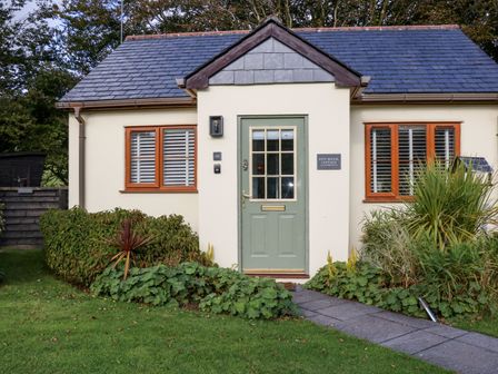 Front view of a cottage with a green door and two windows with shutters surrounded by plants and a grass lawn at Inny Brook Cottage in Davidstow near Camelford