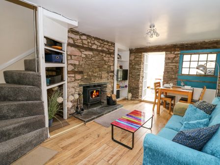 A living room with a blue sofa a colorful coffee table a wood burning stove stone walls and a dining table at Rhif Deg in Brecon