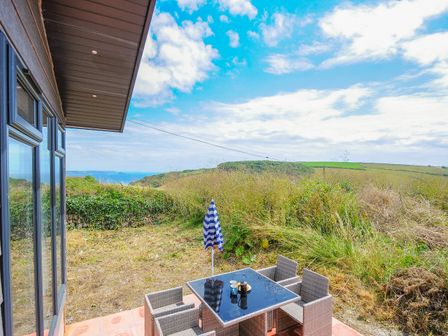 A patio with a table and four chairs next to a house overlooking grassy fields and distant hills at Midway Lodge in Cawsand near Kingsand