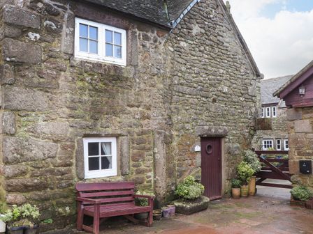A stone cottage exterior with two white windows a maroon door a maroon bench and potted plants at The Old Dairy in Pendeen