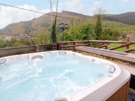 An outdoor hot tub with bubbling water on a wooden deck overlooking a forested hill at Greenheart Cottage in Pontrhydyfen