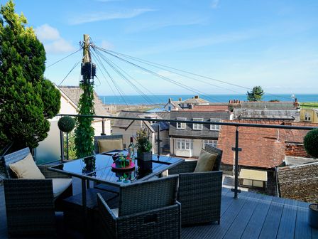 An outdoor balcony with a glass table and four chairs overlooking rooftops and the sea at Lynmouth View in Lynmouth