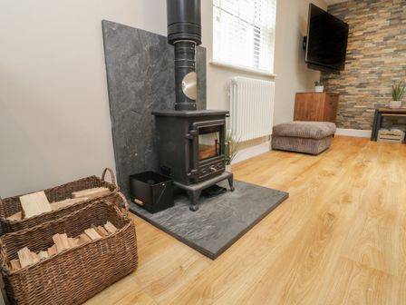 A room with a wood burning stove baskets of firewood a television mounted on a wall and a cushioned stool at Fir Tree Cottage in Llanrug