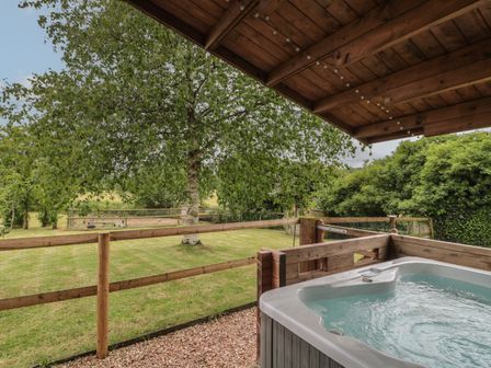 An outdoor hot tub under a wooden roof with a grassy yard and trees at Pigsfoot Lodge in Ash Thomas near Halberton Devon
