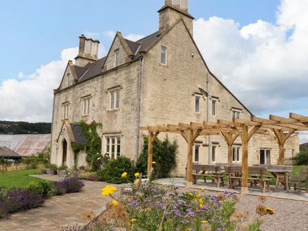 A stone farmhouse with a wooden pergola and outdoor seating surrounded by flowers and greenery at Hammonds Farmhouse in Stroud