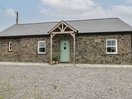 A stone house with two windows a green door and wooden porch on a gravel driveway at Ystabl Robyn in Llanddarog