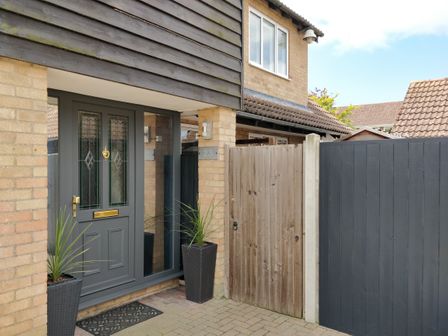 A grey front door with glass panels and a brass mail slot flanked by two tall plant pots next to a wooden gate at 23 Brandeston Close in Great Waldingfield
