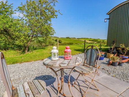 An outdoor patio with a round wooden table and two chairs with a tree and green fields in the background at Puddle Duck Shepherds Hut in Crackington Haven