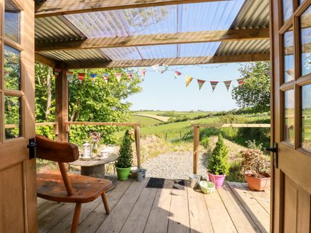 A wooden porch with chair and table surrounded by potted plants and bunting overlooking a rural landscape at Shepherds Retreat in Maxworthy near Crackington Haven