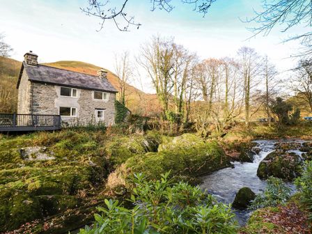 A stone house next to a flowing river with moss-covered rocks and leafless trees near Pen y Bont Near Mallwyd