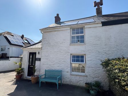 The exterior of a white cottage with two windows a black door and a blue bench with plants in pots outside Blacksmiths Cottage in Crantock