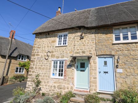 A stone cottage with two light blue doors and white framed windows with flowers outside at Dormouse Cottage in Burton Bradstock