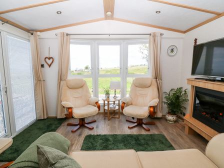 A living room with two beige armchairs facing a window a small round table with a lamp a television a fireplace and green rugs at Gwel Y Mor in Nasareth near Caernarfon
