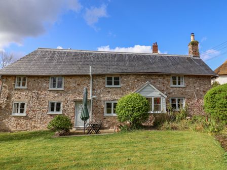 A stone cottage with a slate roof a small patio table and chairs and bushes in the garden at Bridge Cottage in Dalwood