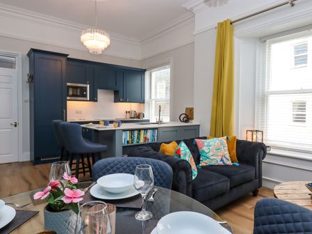 A kitchen and living area with blue cabinets a dark sofa with colorful cushions and a glass dining table with place settings at Sailors Rest in Dartmouth