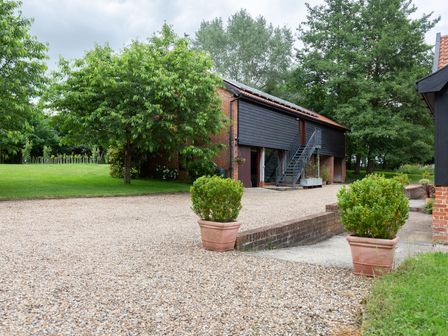 An outdoor area with a gravel driveway two potted plants green trees and a black wooden building at The Cart Lodge at Grove Barn in Thornham Magna near Eye