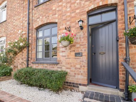 The exterior of a brick house with a dark grey door and window with hanging flower baskets and bushes at Ross Cottage Broadway