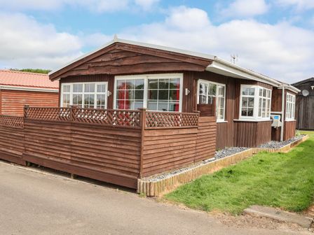 A wooden cottage with a fenced patio on a paved road with a grass lawn at Swiss Cottage in Wilsthorpe near Bridlington