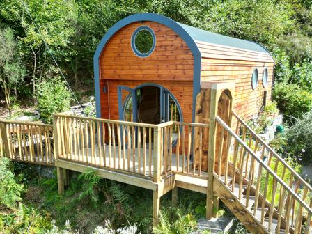A wooden cabin with circular windows and a deck with a railing surrounded by greenery at The Kites Nest in Pont-rhyd-y-groes near Pontrhydfendigaid Snowdonia