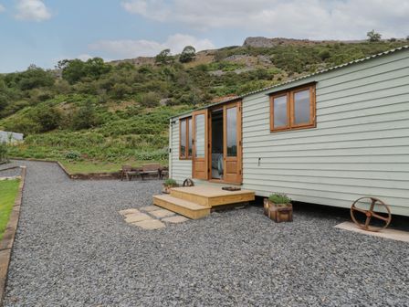 The exterior of a light green wooden shepherds hut with open doors on a gravel surface with a hill in the background at Tan Y Castell Shepherds Hut in Llangollen