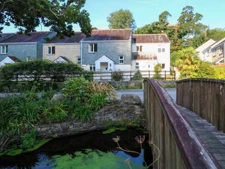 A wooden footbridge over a pond with houses and trees in the background at Primrose Cottage Goldenbank near Falmouth