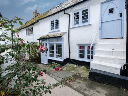 A white house with blue windows and doors with stairs leading to an elevated door and flowers in the foreground at St Samson in Port Isaac