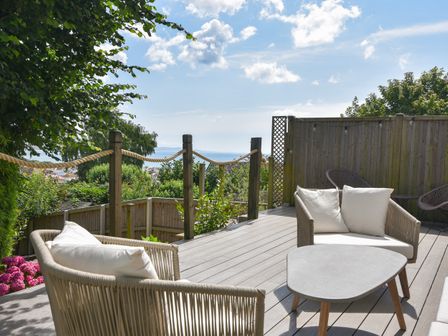 An outdoor deck with seating cushions a table rope railing and a view of greenery and the sea at Coastal View in Lyme Regis