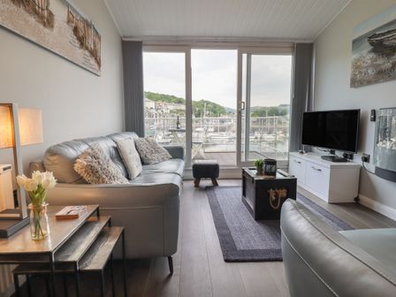 A living room with a gray sofa, coffee table, television, lamp, and a view of boats outside at Moelwyn 57D South Snowdon Wharf in Porthmadog