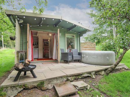 A small green cabin with open double doors revealing an interior with kitchen utensils and a bed a patio with two chairs and a table a portable hot tub and a fire pit at Ty Bach Twt in Talybont-on-Usk
