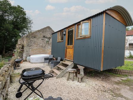 An outdoor area with a corrugated metal shepherd hut a grill two chairs a hot tub and wooden stools at Ty Olwyn Bach in Talybont-on-Usk