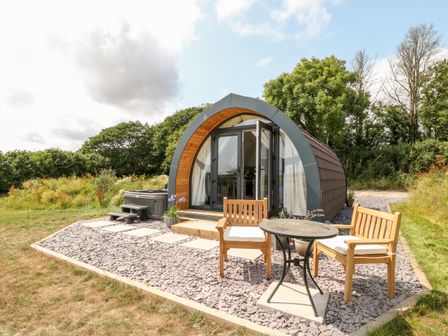 A small cabin with wooden furniture and a table outside on a gravel patio surrounded by grass and trees at Oak in Elmgate near Saltash