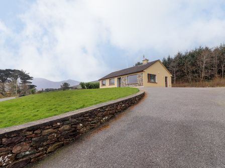 A single-story house with stone and yellow walls on a grassy hill near a paved driveway and trees at Gortnamona in Coolnaharragill Lower near Glenbeigh County Kerry