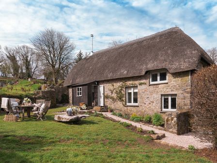 A stone cottage with a thatched roof and outdoor wooden table and chairs set on a grassy lawn at Butterwell Cottage at Collihole in Chagford