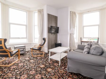 A living room with gray sofa, two armchairs, white coffee table, wall-mounted TV, standing lamp, and large windows at 5 Beach View Beaconsfield House Bridlington