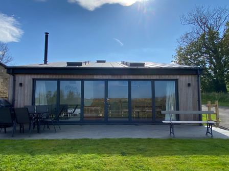 A wooden building with large glass doors a patio table with chairs and a bench on a lawn at 1 Orchard Cottages Dunstan near Craster