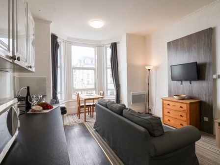 A living room with a sofa a wooden chest of drawers a mounted television and a table with chairs by large windows at 4 Beach View Beaconsfield House in Bridlington