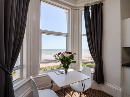 A small dining area with a table flowers in a vase and two chairs by windows with a sea view at 6 Beach View Beaconsfield House in Bridlington