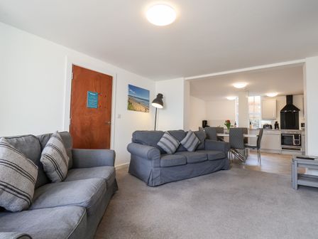 A living room with gray sofas and striped pillows next to a wooden door and an open kitchen with dining table and chairs at Apartment 1 at Bridlington Bay in Bridlington