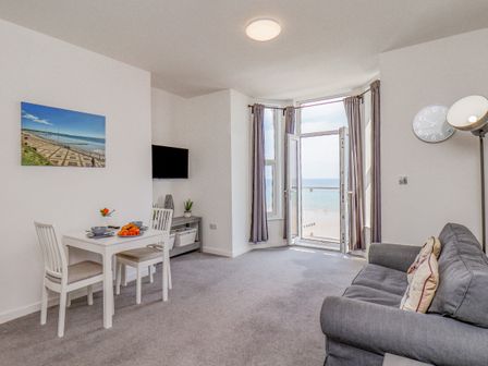 A living room with a dining table and chairs a grey sofa a wall clock and a door opening to a sea view at 4 Seaview Bridlington Bay in Bridlington