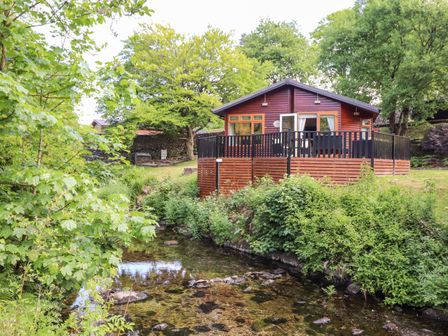 A wooden lodge beside a clear stream surrounded by green trees and shrubs at Nutkins Lodge in Beckside 5 Limefitt Park near Windermere